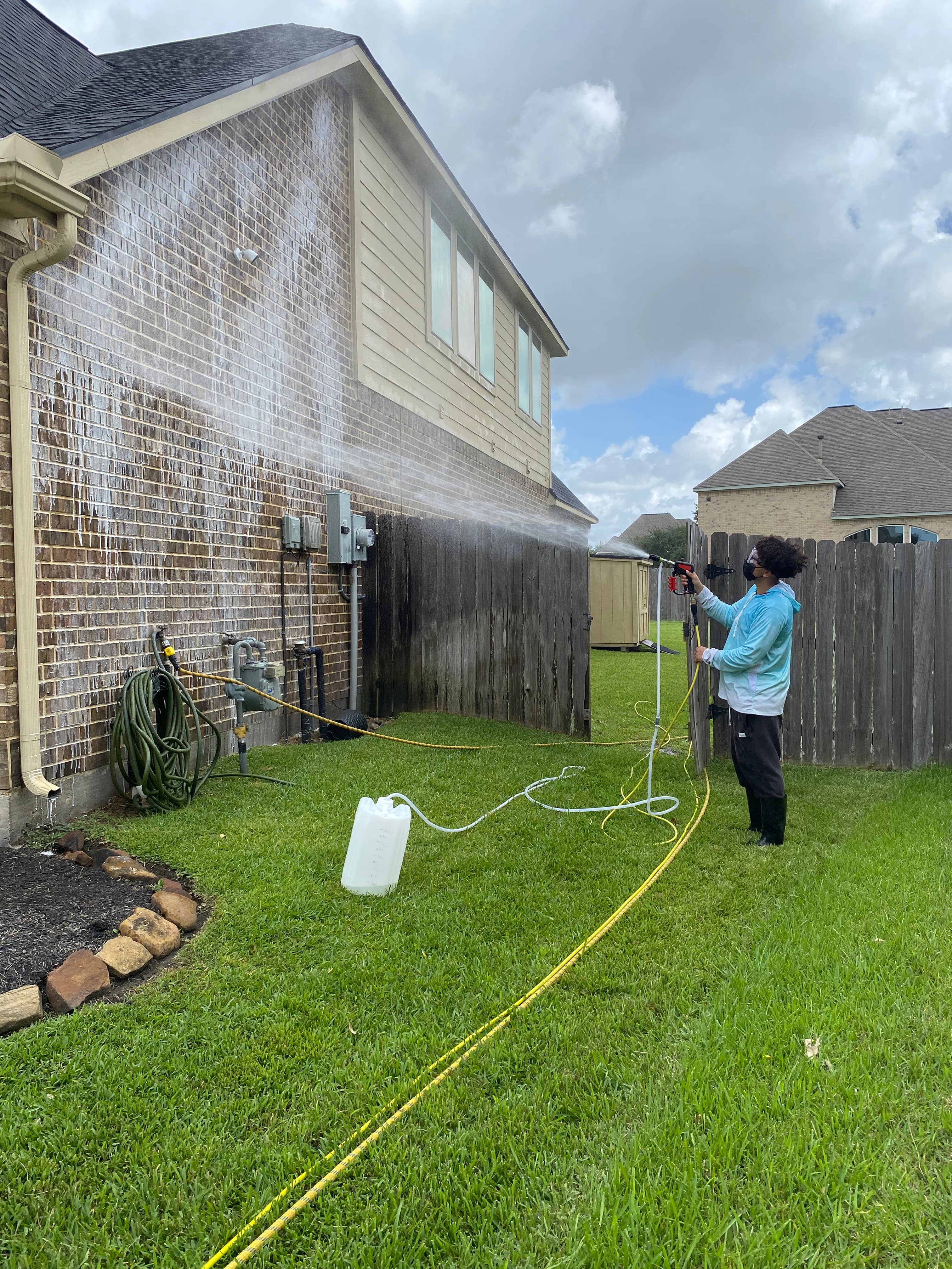 A person in a blue hoodie pressure washes the brick wall of a suburban house.