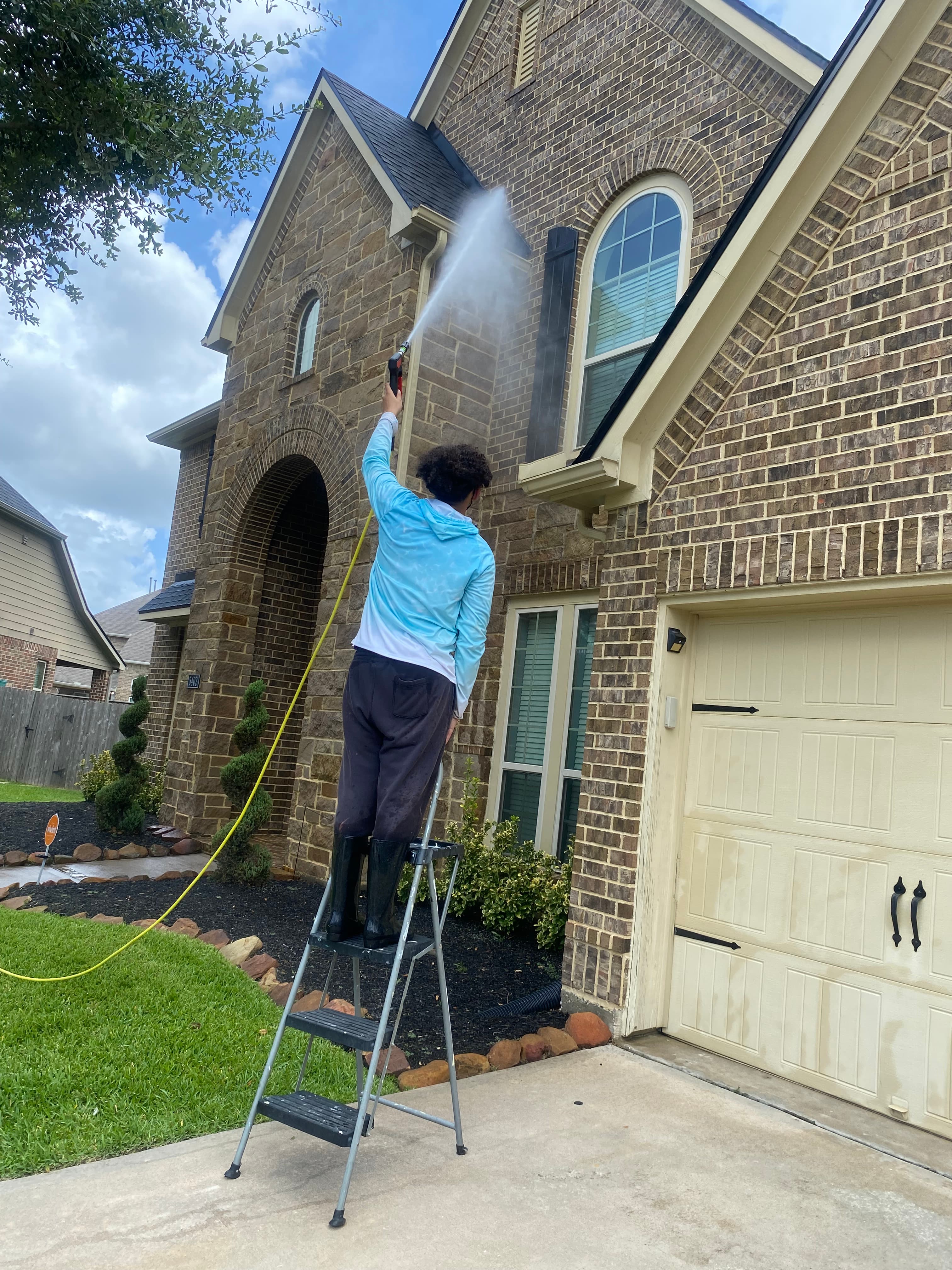Person on a ladder pressure washing the brick exterior of a large suburban house.